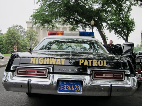 1991 Ford LTD Crown Victoria - Rear of a black Dodge cruiser lettered HIGHWAY PATROL in gold with a California 834428 plate and roof light bar, an officer standing alongs…