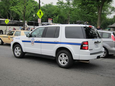 1991 Ford LTD Crown Victoria - Profile of a white Ford Explorer police SUV with a blue stripe and roof equipment, parked beside other cars near a tour bus and pedestrian…
