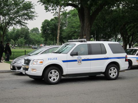 1991 Ford LTD Crown Victoria - Front three-quarter view of a white Ford Explorer marked POLICE with a blue stripe and roof light bar, parked at a curb beside a silver sed…