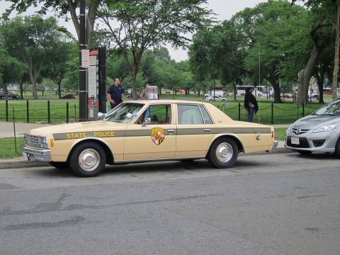 1991 Ford LTD Crown Victoria - Side view of a tan and brown STATE POLICE sedan with a Maryland shield emblem and chrome hubcaps, parked curbside in a tree-filled park.