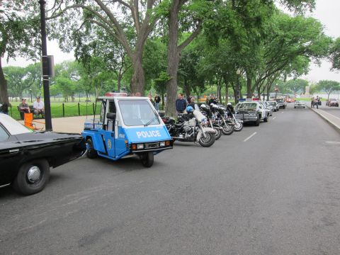 1991 Ford LTD Crown Victoria - blue and white three-wheeled POLICE scooter parked at the head of a row of police motorcycles and cruisers along a tree-lined park road.