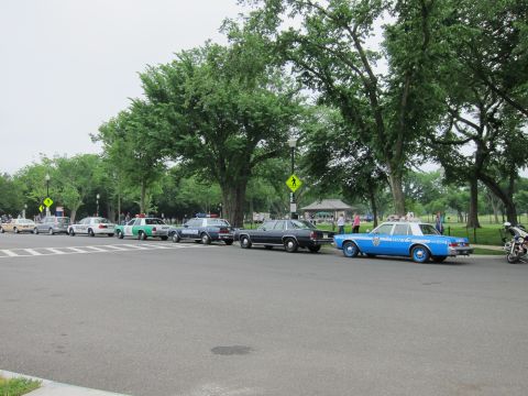 1991 Ford LTD Crown Victoria - lineup of vintage police cruisers parked along a tree-lined park road, with a blue and white NYPD-style sedan and a motorcycle at the right…