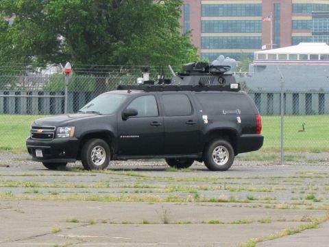1991 Ford LTD Crown Victoria - black Chevrolet Suburban SUV fitted with roof-mounted surveillance equipment, parked in a cracked gravel lot in front of an office building.