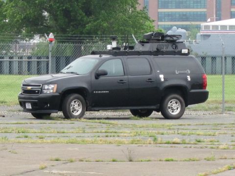1991 Ford LTD Crown Victoria - Profile of a black Chevrolet Suburban with roof-mounted equipment and small markings, parked in a weedy cracked lot before a brick building.