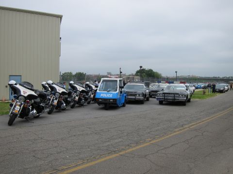 1991 Ford LTD Crown Victoria - lineup of police motorcycles, a blue three-wheeled POLICE scooter, and vintage cruisers parked along a metal building under an overcast sky.
