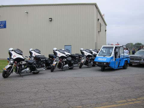 1991 Ford LTD Crown Victoria - Several white police motorcycles lined up next to a blue and white NYPD three-wheeled scooter beside a tan metal building.