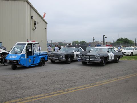 1991 Ford LTD Crown Victoria - blue NYPD three-wheeled scooter parked beside black vintage police sedans and a black-and-white cruiser in a paved lot by a metal building.