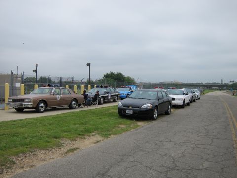 1991 Ford LTD Crown Victoria - long line of police cars stretching from a vintage tan sedan to modern black and white cruisers parked along a paved waterfront path.