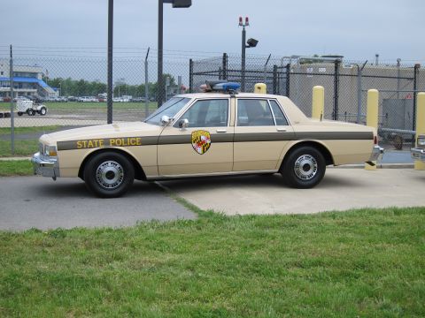 1991 Ford LTD Crown Victoria - Front three-quarter view of a tan and brown STATE POLICE Chevrolet Caprice with a Maryland shield emblem and roof beacons, parked near a ch…