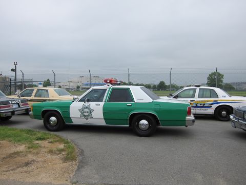 1991 Ford LTD Crown Victoria - Profile of a green and white police sedan with a seven-point star emblem and roof beacons, flanked by a tan sedan and a modern white police…