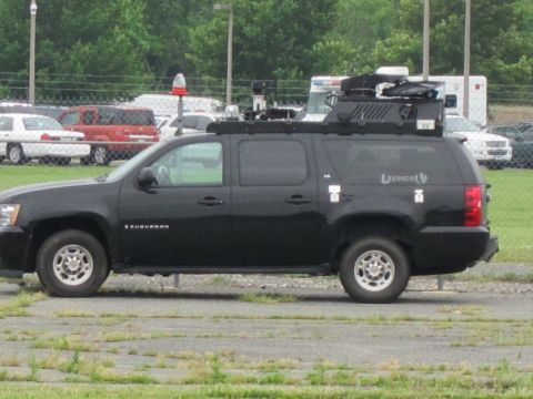 1991 Ford LTD Crown Victoria - black Chevrolet Suburban outfitted with roof-mounted tactical equipment sits in a paved lot beside a chain-link fence.