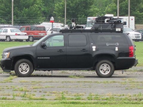 1991 Ford LTD Crown Victoria - Profile of a black Chevrolet Suburban fitted with roof-mounted equipment, parked on pavement near a chain-link fence.