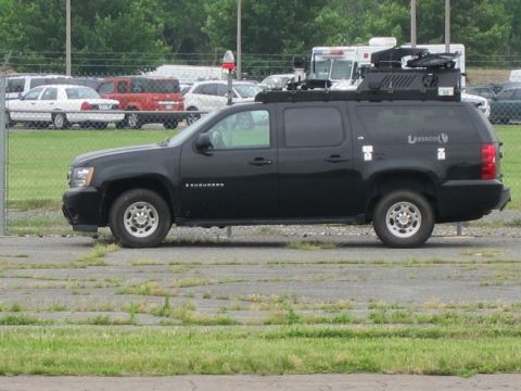 1991 Ford LTD Crown Victoria - black Chevrolet Suburban carrying roof-mounted equipment idles in a lot with a person seated inside.