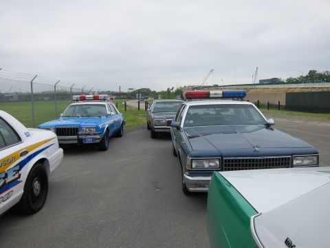 1991 Ford LTD Crown Victoria - group of classic police cars with roof light bars parked on tarmac under an overcast sky near a fence.