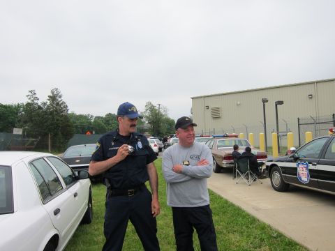 1991 Ford LTD Crown Victoria - uniformed officer and a man in a ball cap converse beside parked white police cars near a metal building.