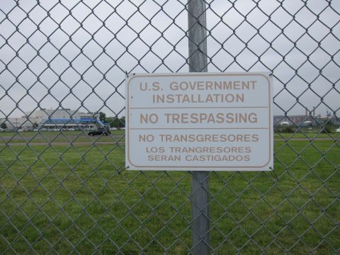 1991 Ford LTD Crown Victoria - white sign reading U.S. Government Installation No Trespassing is mounted on a chain-link fence over a grassy field.