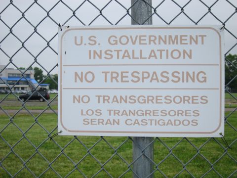 1991 Ford LTD Crown Victoria - No Trespassing sign for a U.S. Government Installation hangs on a chain-link fence with a dark car and building beyond.