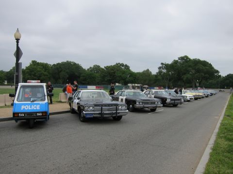 1991 Ford LTD Crown Victoria - long line of vintage police cars and a small three-wheeled police cart are parked along a roadside under a cloudy sky.
