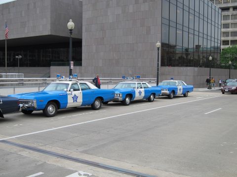 1991 Ford LTD Crown Victoria - Three blue and white Chicago police cars with star emblems are parked along a city street beside a large stone building.
