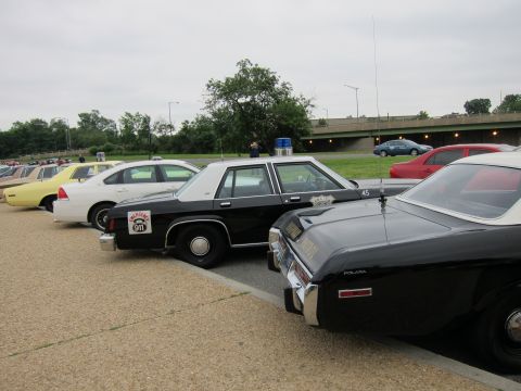 1991 Ford LTD Crown Victoria - Black-and-white vintage police cars, including a Plymouth and a 911 sticker car, lined up near a grassy highway embankment.