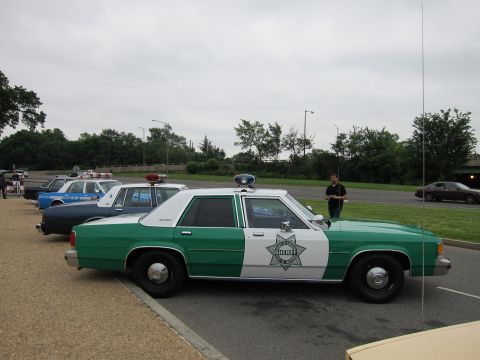 1991 Ford LTD Crown Victoria - Side profile of a green-and-white Ford sheriff cruiser parked with other police cars, a man standing nearby.