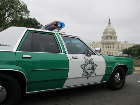 1991 Ford LTD Crown Victoria - Rear quarter of a green-and-white sheriff cruiser with star decal, the US Capitol dome visible in the background.