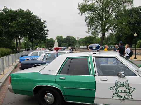 1991 Ford LTD Crown Victoria - Green-and-white sheriff cruisers with California county star decals parked along a fenced path, people standing nearby.