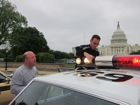 1991 Ford LTD Crown Victoria - man reaches over the roof of a white police car to adjust its lightbar, the US Capitol dome behind.