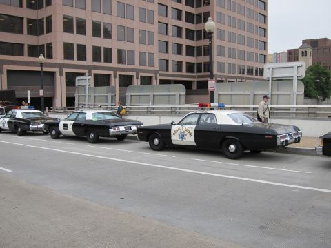 1991 Ford LTD Crown Victoria - line of black and white vintage police cars, including a California Highway Patrol cruiser, parked along a downtown street by office buildi…