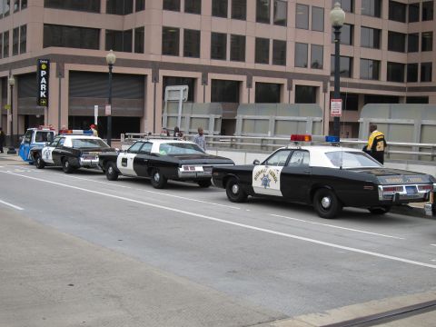 1991 Ford LTD Crown Victoria - Several black and white vintage police sedans with roof light bars, including a Highway Patrol cruiser, line a downtown curb near a parking…