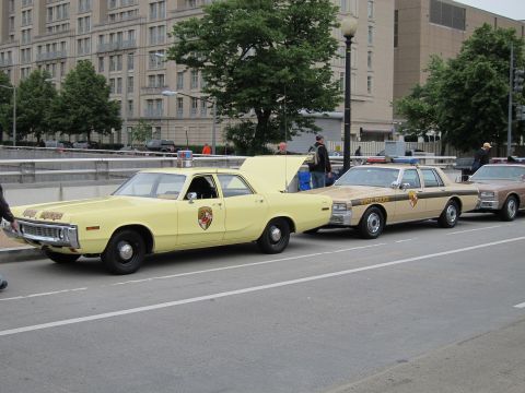 1991 Ford LTD Crown Victoria - pale yellow vintage state police sedan with a round crest emblem is parked along a city curb ahead of other classic cruisers.