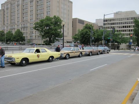 1991 Ford LTD Crown Victoria - pale yellow vintage state police sedan with a crest emblem heads a line of classic police cruisers parked along a wide city street.