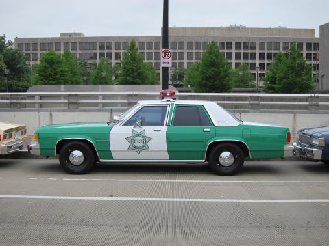 1991 Ford LTD Crown Victoria - green and white Ford sheriff sedan with a star emblem and single roof beacon is shown in side profile parked along a city curb.