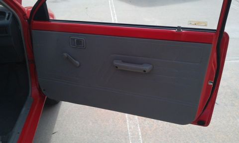 1991 Ford Festiva - The interior door panel of a red vehicle shows a gray trim with a manual window crank and a pull handle.
