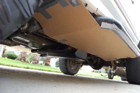 1994 Ford Bronco - Ground-level view under a truck's rocker area where large cardboard panels have been duct-taped along the body and floor as masking for und…