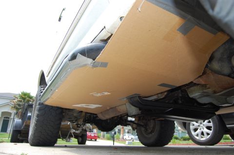 1994 Ford Bronco - Low ground-level view under the rocker of a silver truck in a driveway, where large cardboard sheets have been duct-taped along the body si…