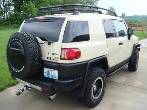 2010 Toyota FJ Cruiser - This tan Toyota FJ Cruiser is seen from the rear, a spare tire on the back and a roof rack. The FJ Cruiser badge is visible on the tailgate.