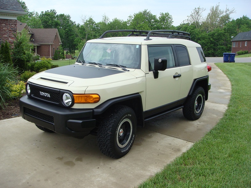 Tan Toyota FJ Cruiser parked on a driveway, front grille has TOYOTA text. Roof rack installed
