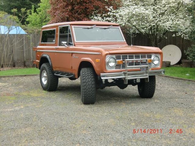 1973 Ford Bronco parked on gravel with trees and a white flowering bush in the background. License plate reads '1973 BRONCO'