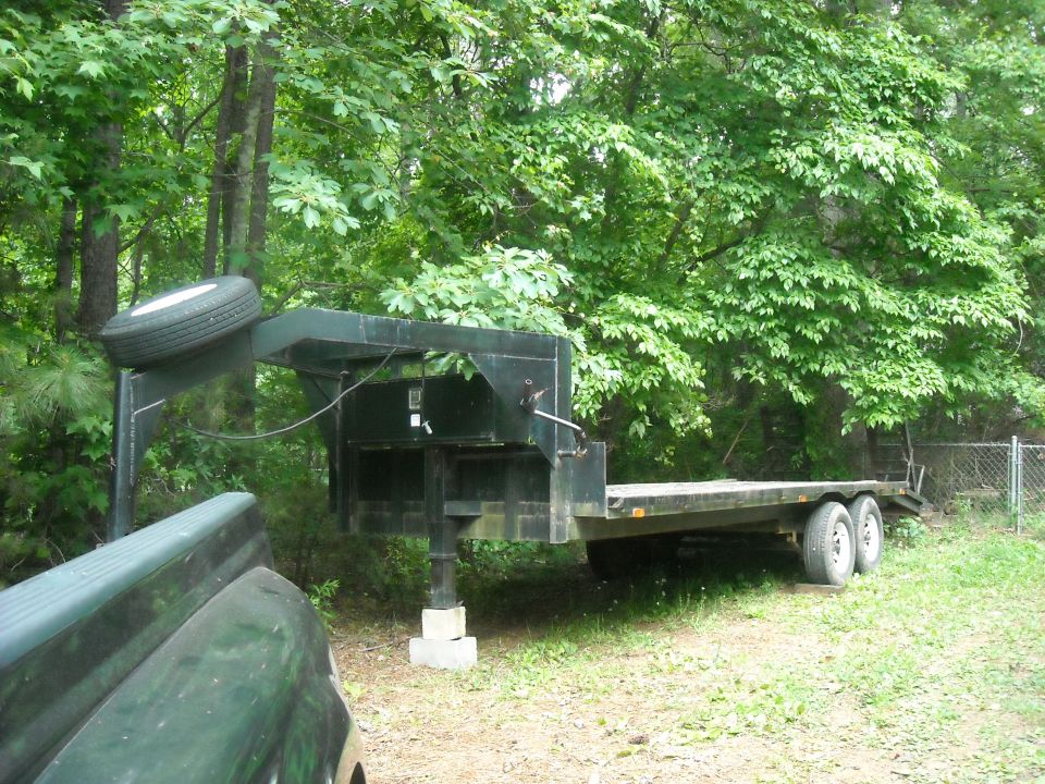 Gooseneck trailer parked in grassy area with trees and fence in background. Spare tire mounted on front. Part of a dark green truck visible on left