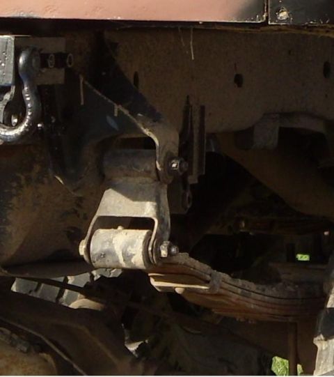 1988 Ford Bronco - Close-up under a truck frame rail showing a leaf spring hanger with a reversed (flipped) shackle bolted to the frame bracket, the leaf pack…