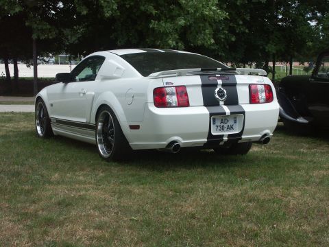1977 Pontiac Trans Am - White 2005 Ford Mustang GT with black stripes and custom wheels sits on a grassy lawn next to another vehicle.