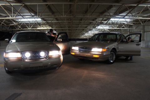 2000 Mercury Grand Marquis - Two Mercury Grand Marquis sedans are parked indoors, with their doors open and headlights illuminated.