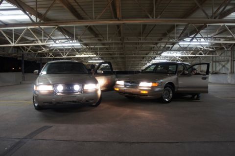 2000 Mercury Grand Marquis - Two tan 2000 Mercury Grand Marquis sedans are parked indoors, one with its door open, showing their front ends.