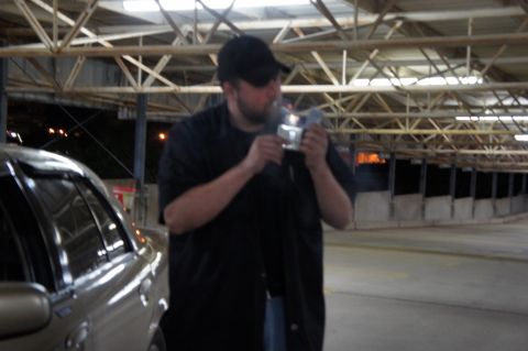 2000 Mercury Grand Marquis - A man lights a cigarette next to a gold 2000 Mercury Grand Marquis sedan parked under a covered structure.