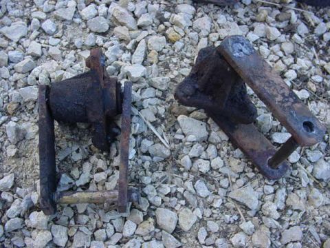 1992 Ford Bronco - Two heavily rusted metal suspension components lie on a bed of gravel, suggesting they are replacement parts.