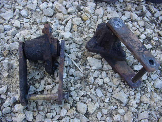 Two rusty metal suspension parts on gravel, one with a cylindrical component