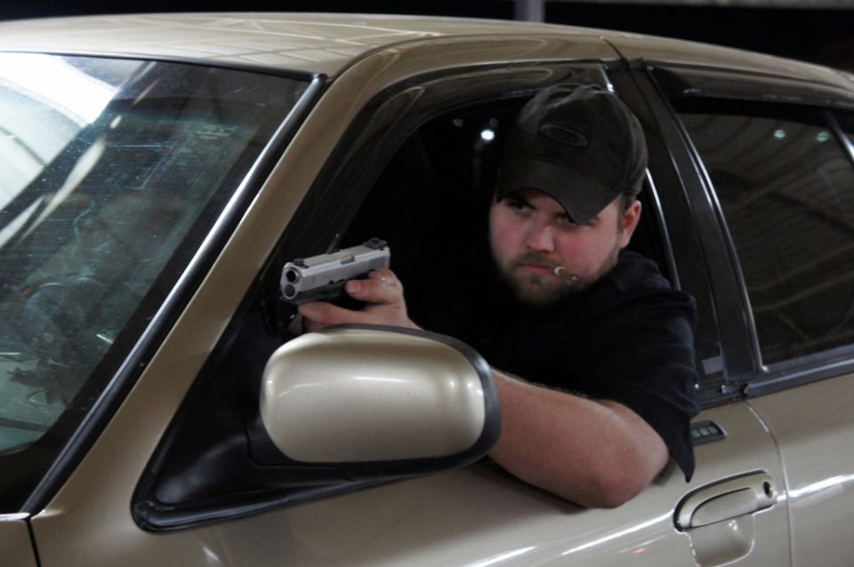 Guy in a black cap and shirt leans out car window holding a pistol, cigar in mouth