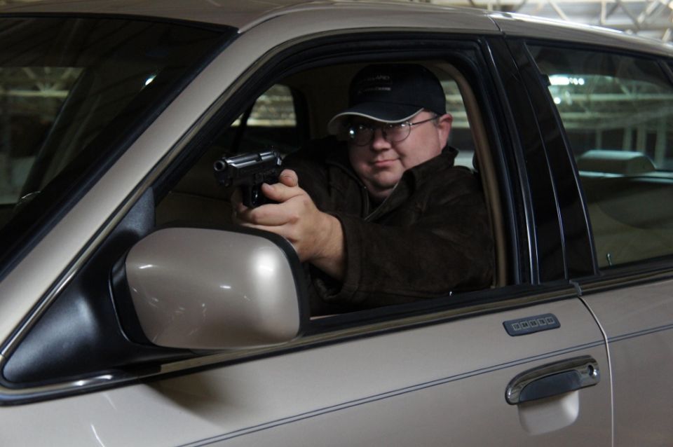 Man in a cap and glasses holding a handgun while sitting in the driver's seat of a 2000 Mercury Grand Marquis