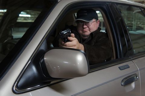 2000 Mercury Grand Marquis - A man wearing a baseball cap and glasses points a handgun while sitting in the driver's seat of a car.